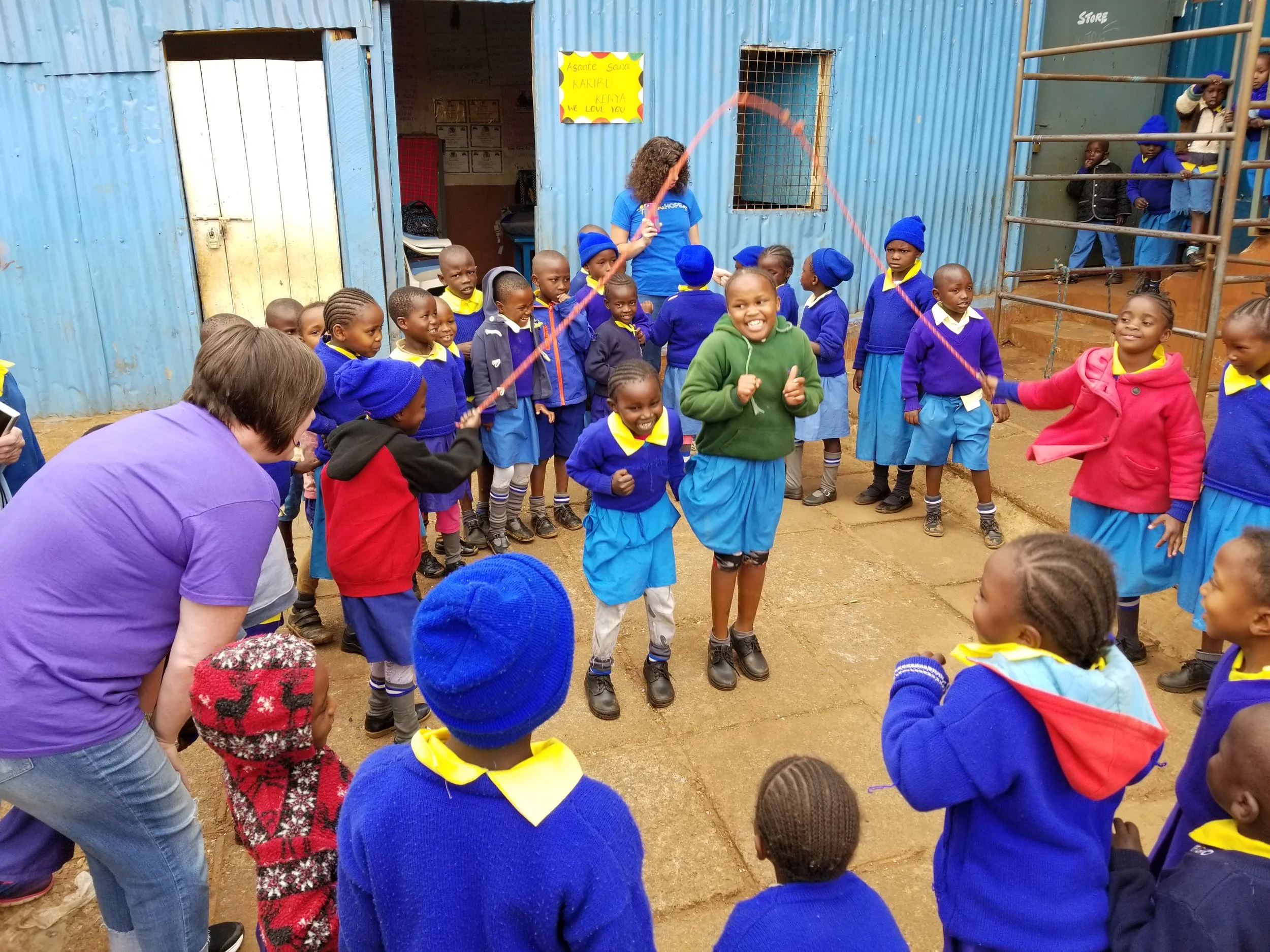Children in school uniforms playing jump rope outside a blue metal building.