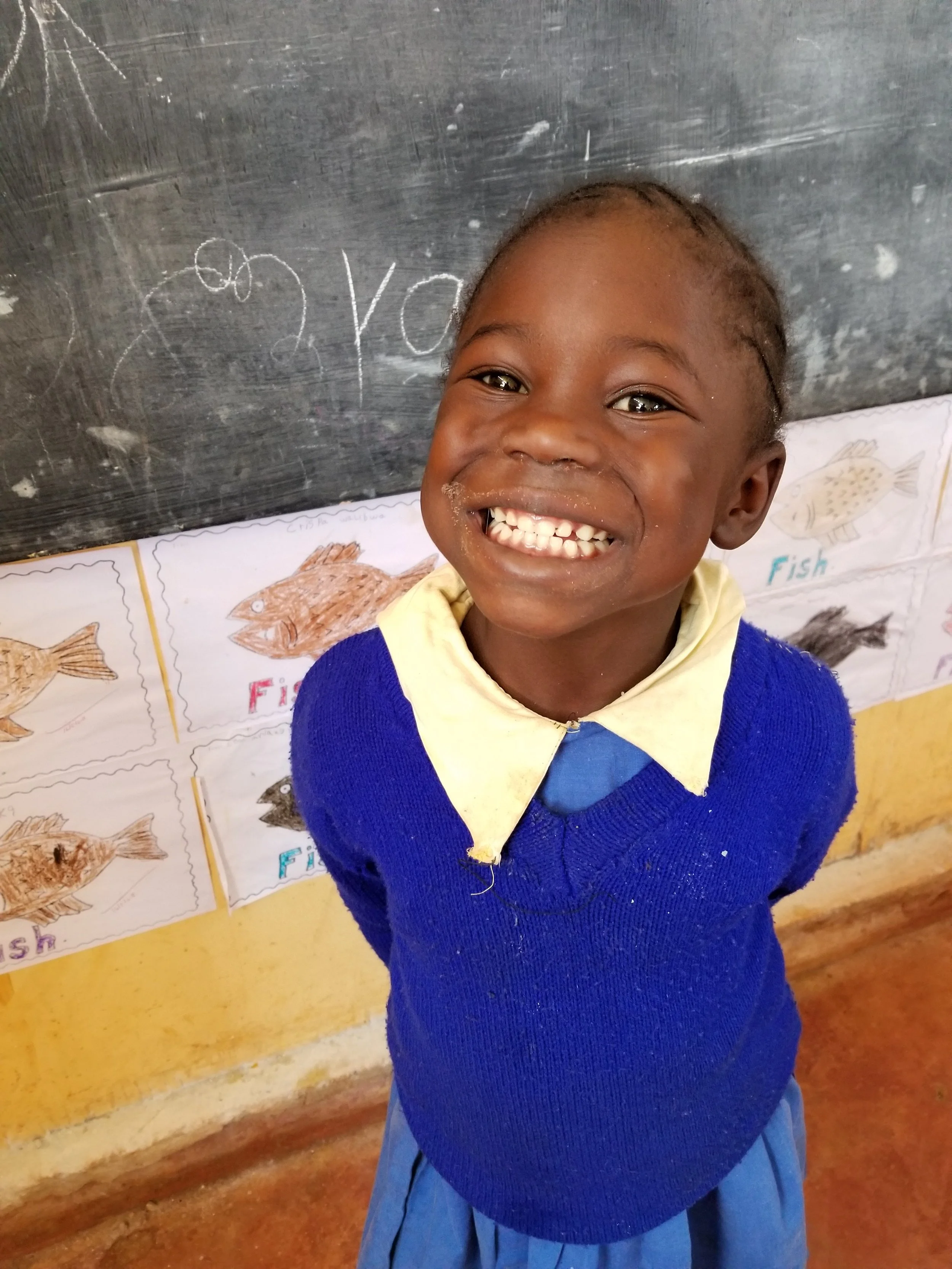 A young girl smiling widely at the camera, wearing a blue sweater and yellow collared shirt, standing in front of illustrated posters of fish on a classroom wall.