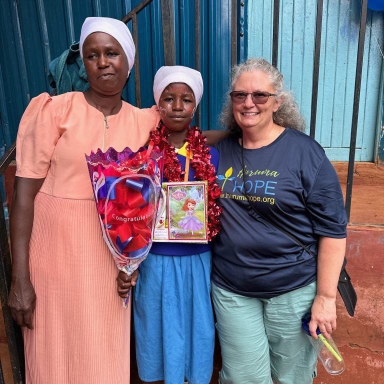 Three women standing together, one holding a bouquet of flowers, another with a medal and decorations, and the third wearing a 'Huruma Hope' shirt, smiling in front of a blue fence.