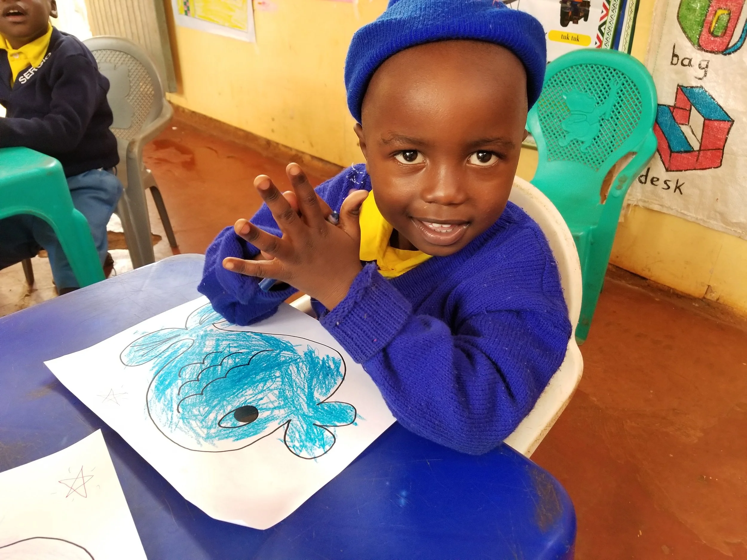 A young boy wearing a blue beanie and blue sweater sitting at a table with a drawing of a dinosaur on paper, smiling at the camera in a classroom.