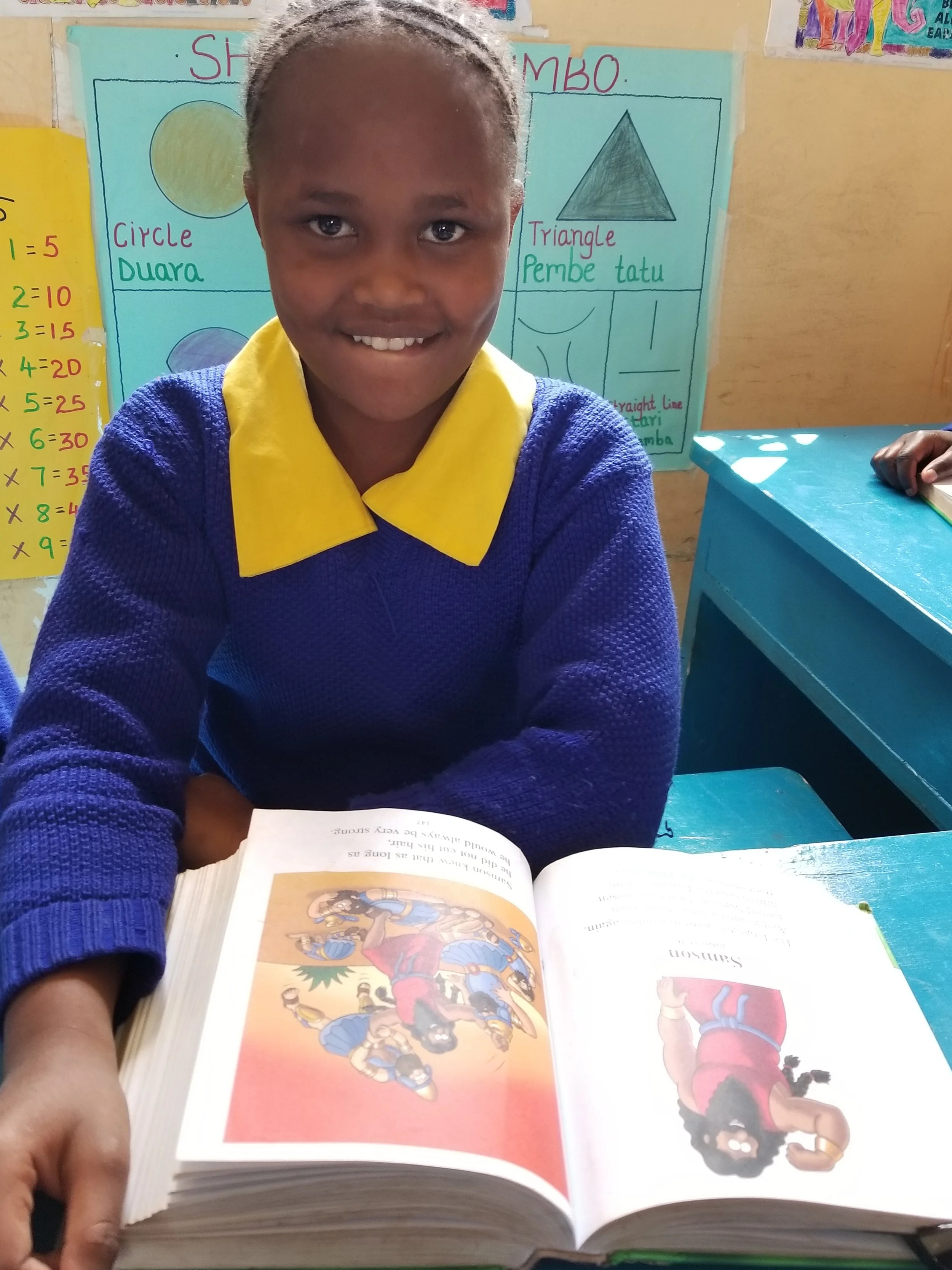 A young girl with a bright smile sitting at a blue desk in a classroom, holding an open book with colorful illustrations. She is wearing a blue sweater with a yellow collar. Behind her are educational posters about shapes and multiplication tables.