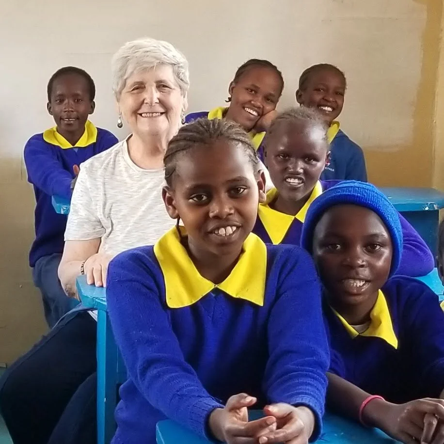 A group of school children in matching blue and yellow uniforms with a smiling elderly woman in casual clothes in a classroom.
