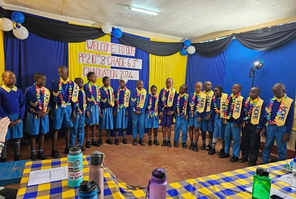 Group of children in blue school uniforms and colorful leis standing on stage for a graduation ceremony with a backdrop that reads 'Welcome to our PP2 10th & Grade 6 3rd Graduation 2024' in a decorated room with balloons.