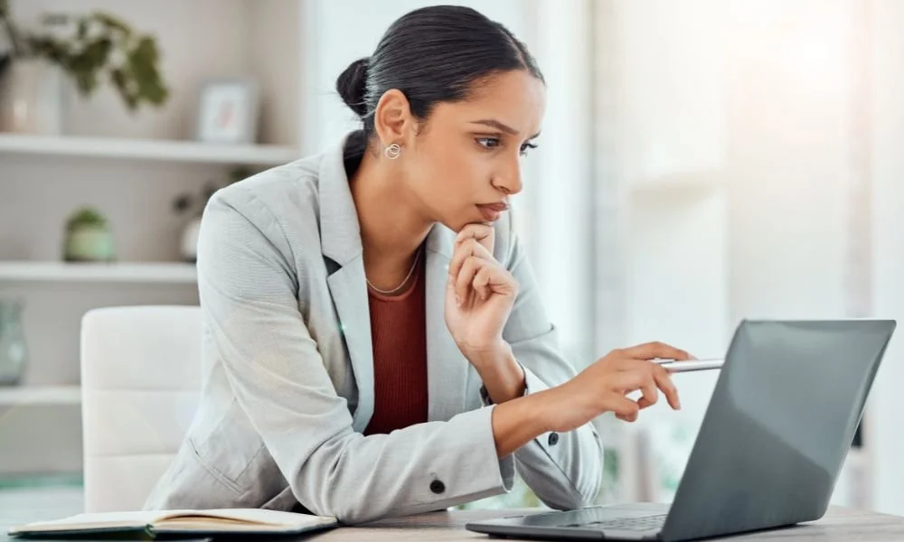 Businesswoman focusing on her laptop while sitting at a desk with a book, in a well-lit office.