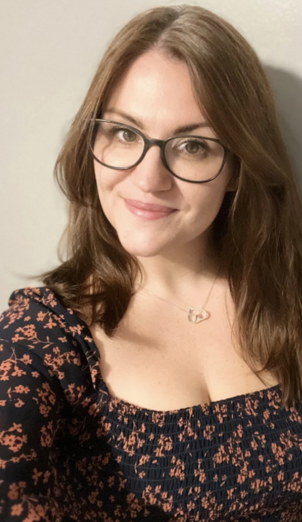 A woman with shoulder-length brown hair, wearing glasses, a floral patterned top, and a heart-shaped necklace, smiling at the camera.