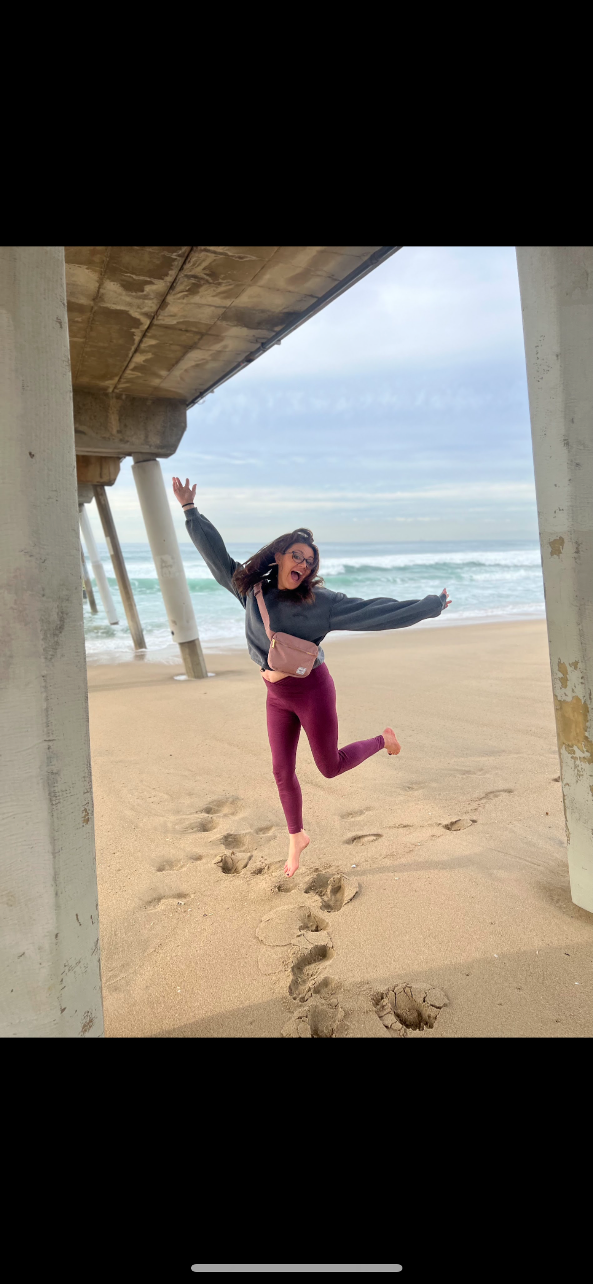 A woman jumping joyfully on the beach under a pier, with sand footprints behind her, ocean waves in the background, wearing glasses, a black jacket, and purple leggings.