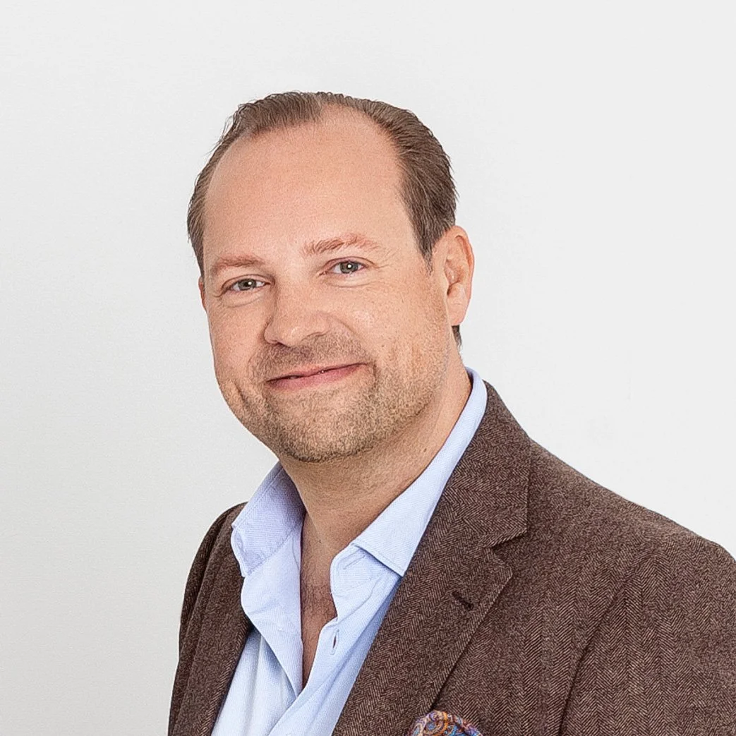 Der Geschäftsführer Hospitality, David Nünemann, in einem braunen Anzug und weißem Hemd, lächelnd gegen einen weißen Hintergrund.