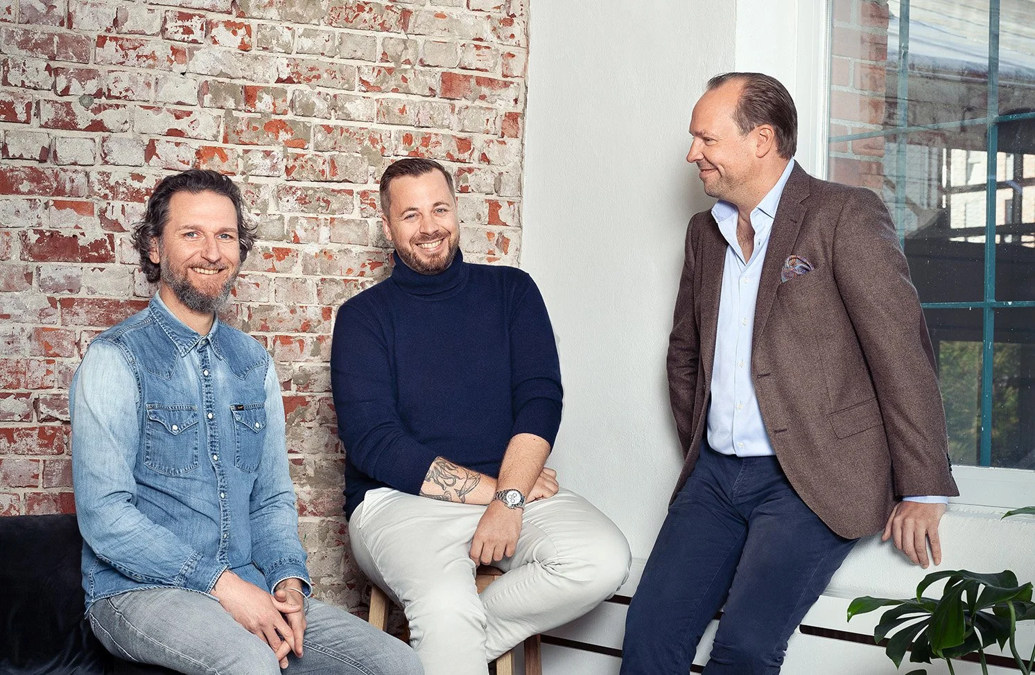 Drei Männer unterhalten sich in einem modernen Büro mit einem roten Backsteinwand und großen Fenstern.
