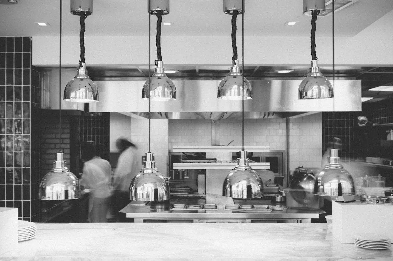 Kücheninnenseite in einem Restaurant mit vier hängenden Lampen und zwei vom Küchenpersonal in Bewegung.