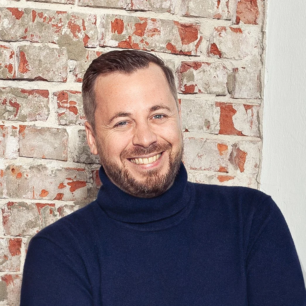 Ein lachender Mann mit kurzhaarigem dunkelblondem Haar, Bart und blauen Augen trägt einen dunkelblauen Rollkragenpullover, steht vor einer alten Ziegelwand.