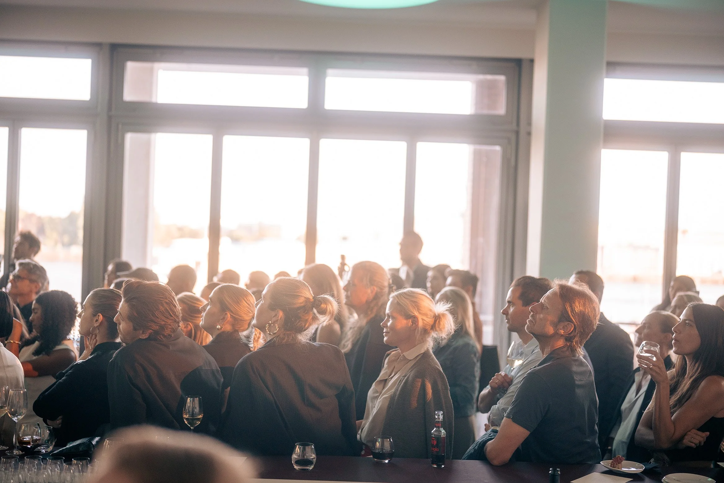 Menschen, die bei einem Event, Launch von der neuen JIL SANDER Kollektion, an einem Tisch sitzen und zuhören, mit Getränken in der Hand, im Hintergrund große Fenster mit Blick auf die Stadt bei Tageslicht. Credit: Andreas Behr.