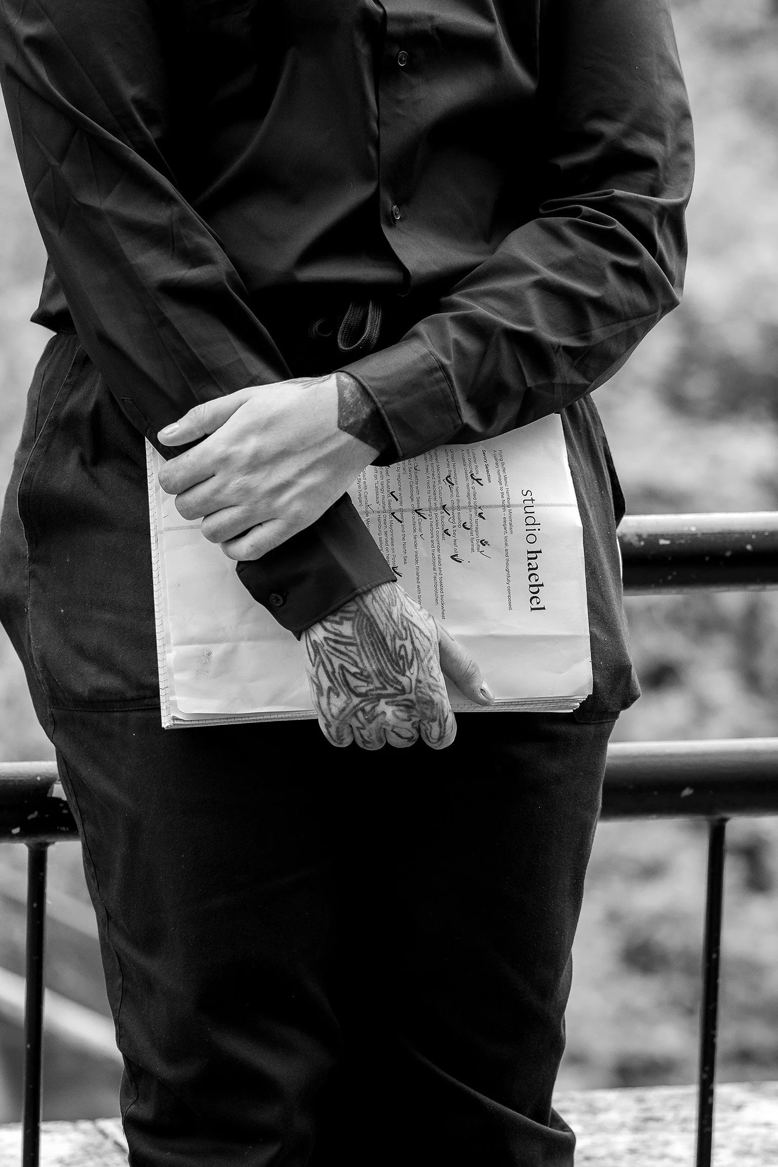 Person in schwarzer Kleidung hält ein Notizbuch mit dem Titel 'studio habel' in der Hand, die Person hat sichtbare Hand- und Armsleeves sowie eine Tätowierung auf der Hand. Im Hintergrund ist eine Metallbarriere zu sehen.