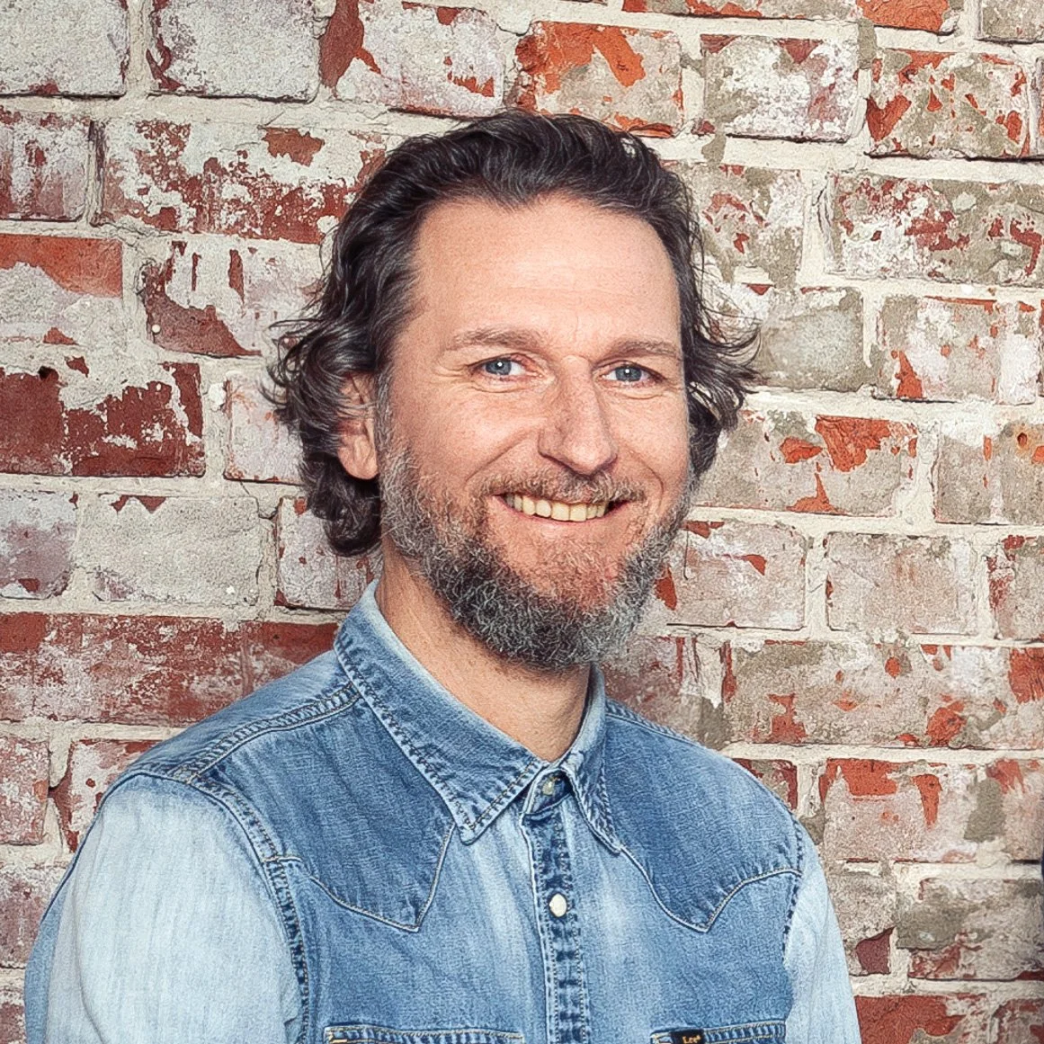 Ein Mann, Nico Lee Gogol, mit dunklen, lockigen Haaren und Vollbart, lächelnd, trägt ein blauen Jeanshemd, steht vor einer alten, gemauerten Wand.