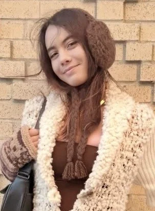 Young woman with long brown hair wearing brown earmuffs, a fuzzy beige sweater, and a brown top, standing in front of a brick wall.