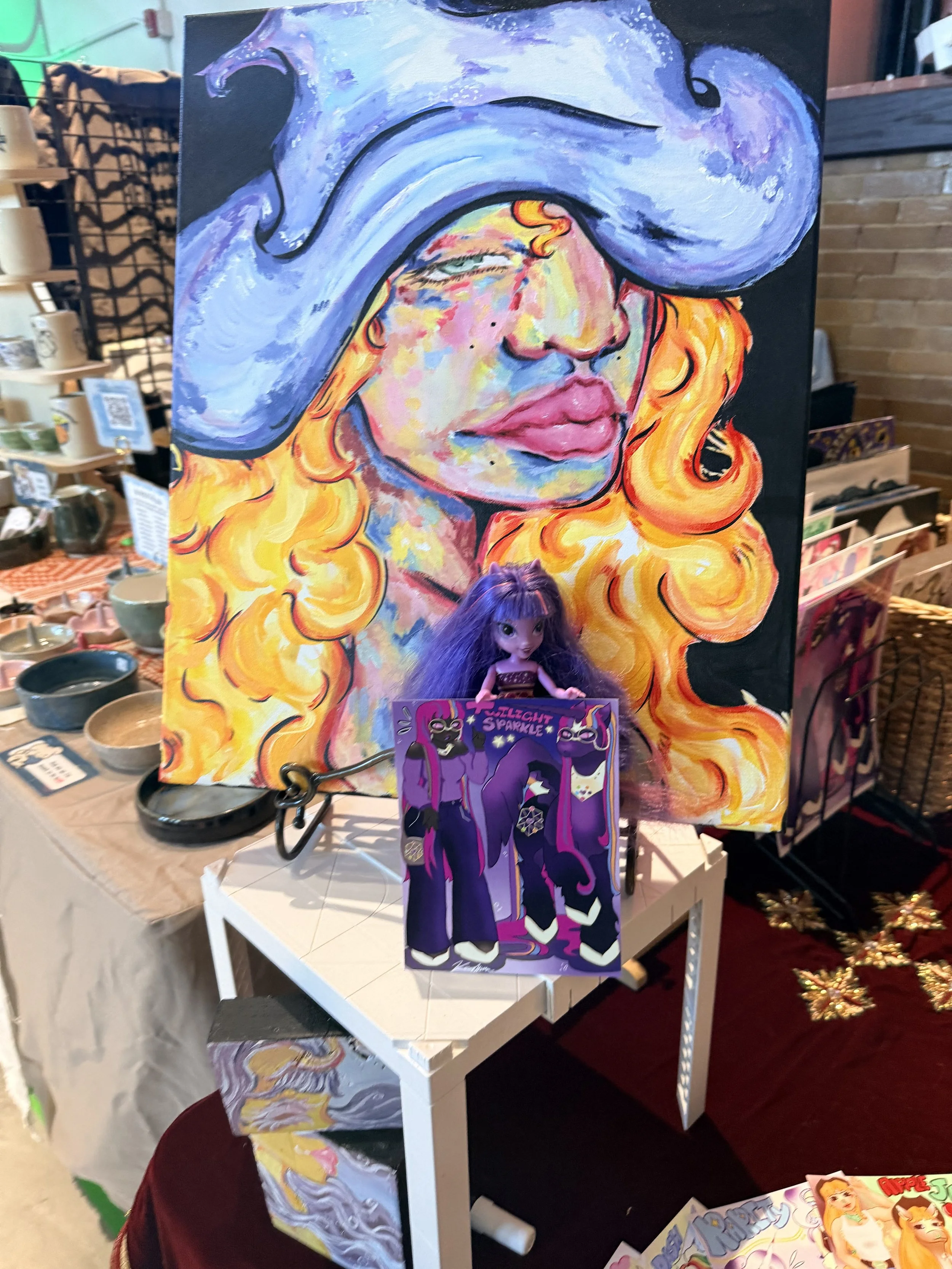 A colorful painting of a woman with long, curly orange hair wearing a large purple hat with a brim that covers her eyes. The painting is on a black background and displayed on an easel. There are various small bowls and items on a table around the painting, and other artwork is visible on shelves and nearby tables in a shop setting.