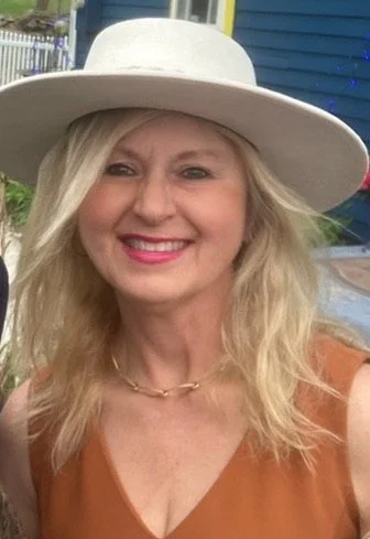 A woman smiling outdoors, wearing a large white sunhat, a brown sleeveless top, and a gold necklace.