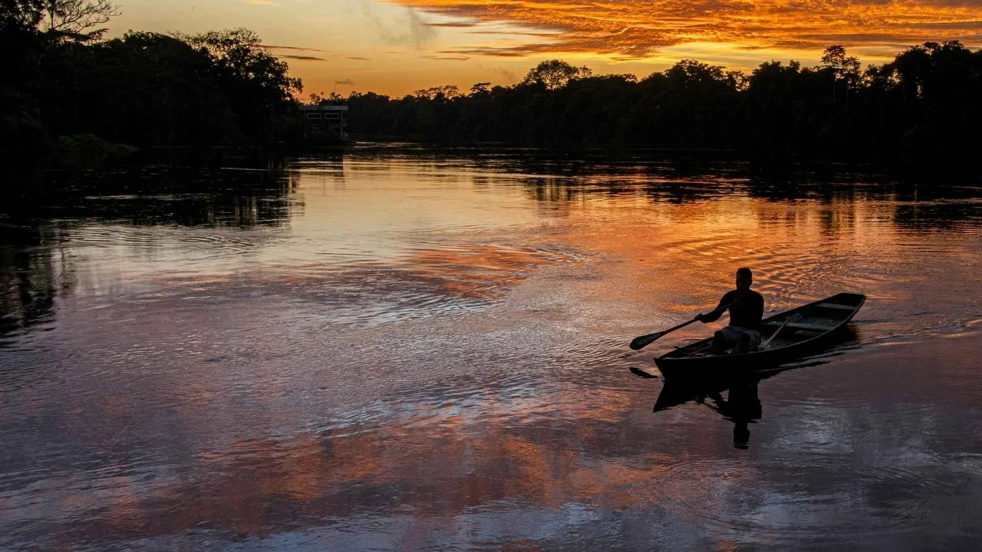 Homem remando um barco em um rio ao pôr do sol, com o céu laranja e refletido na água.