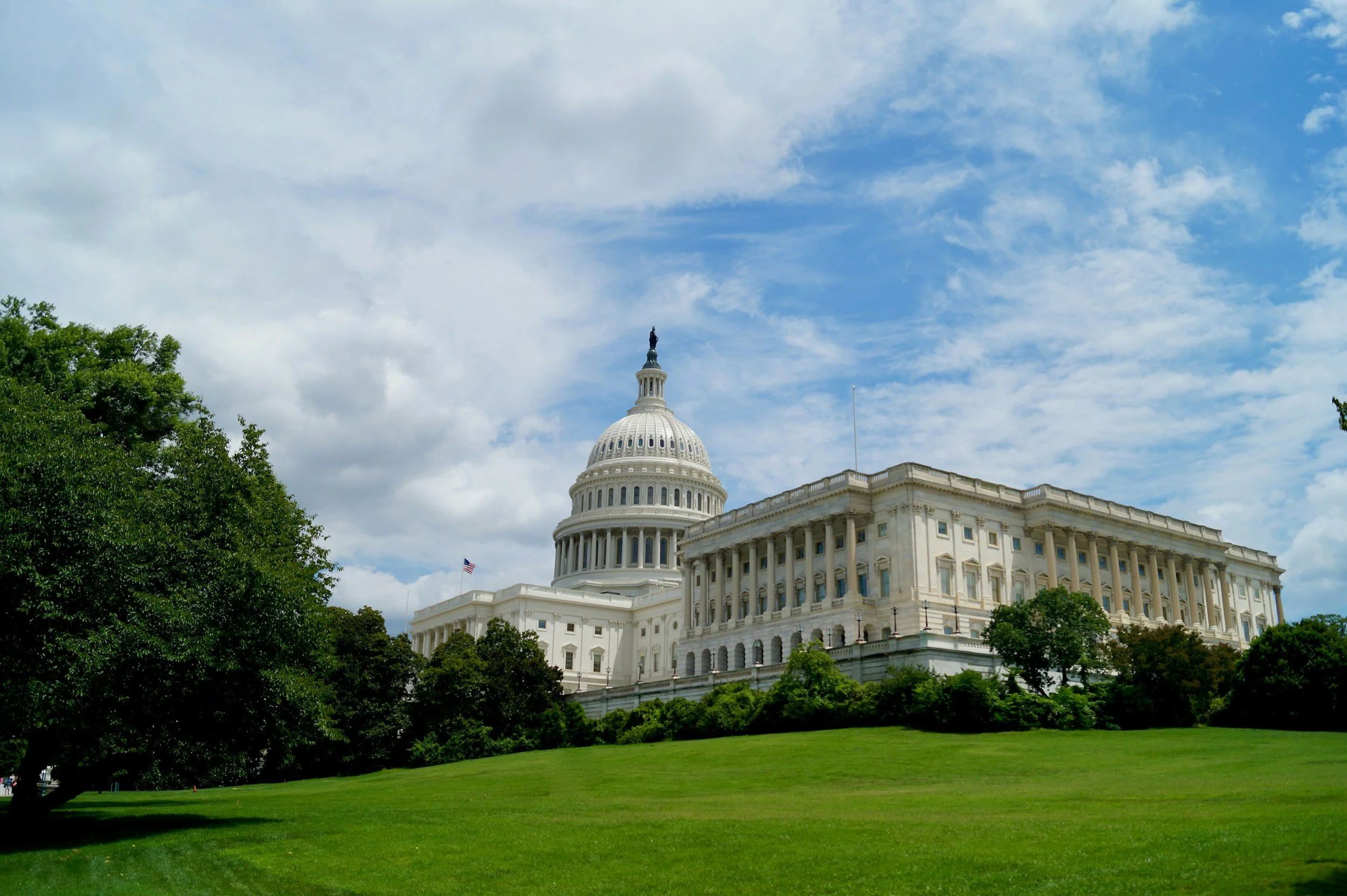 U.S. Capitol building viewed from a distance with a foreground of green grass and trees, partially cloudy sky.