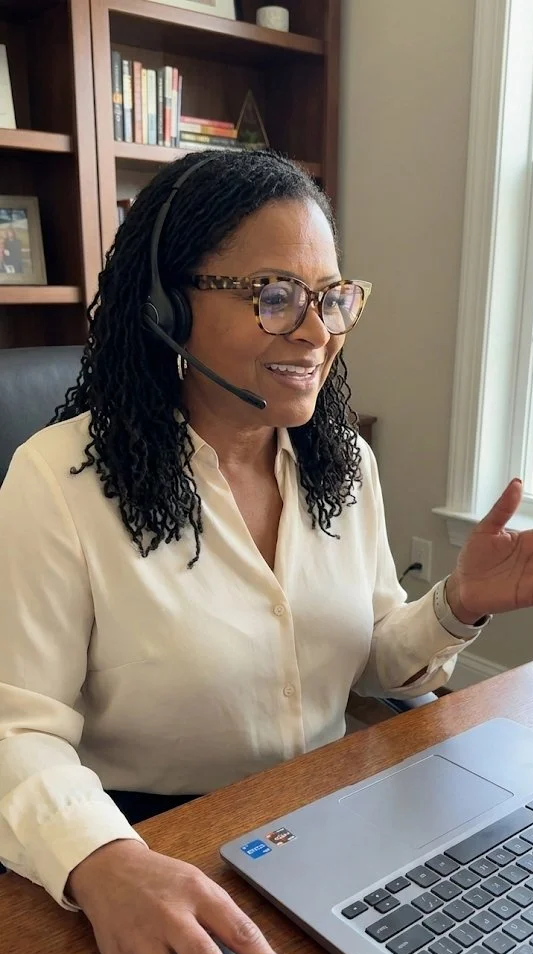 Woman with glasses and a headset smiling and gesturing while working at a desk with a laptop, in a home office with bookshelves in the background.