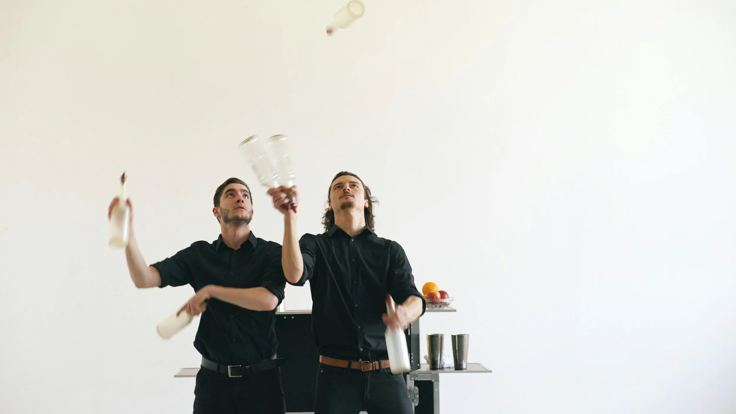 Two men in black shirts juggling bottles and glasses in a minimalist white room.
