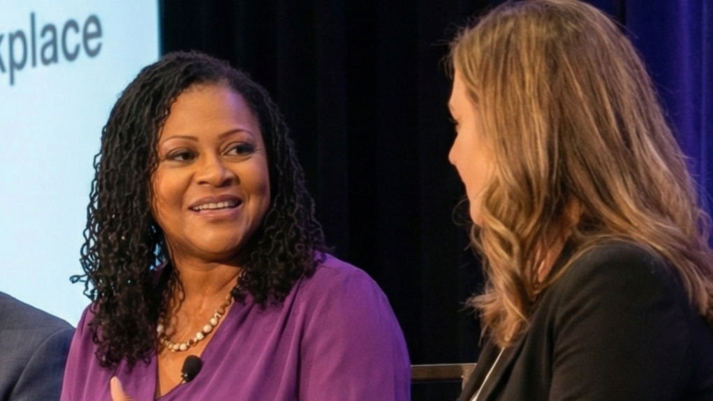 Two women having a conversation on stage at an event, with one woman wearing a purple top and the other with a black blazer, in front of a presentation screen.