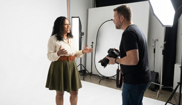 A woman is speaking and gesturing with her hands in a photo studio, while a man is taking photos with a camera.