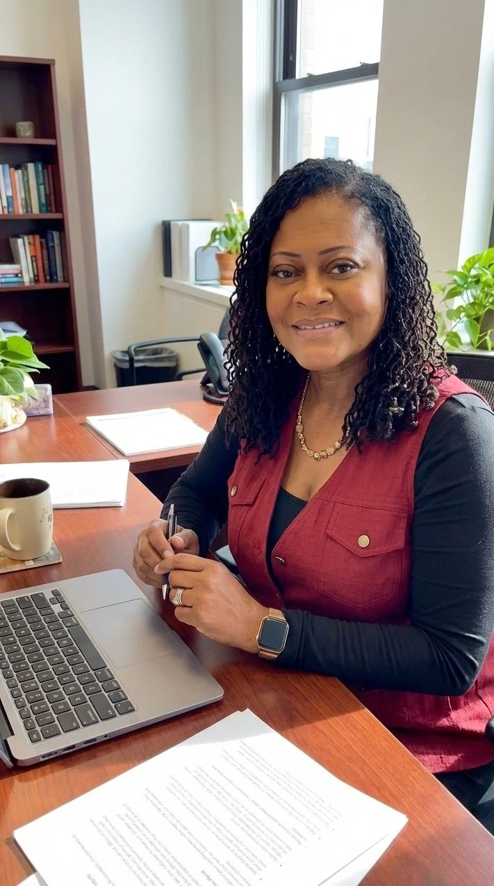 A woman with curly black hair smiling at the camera, sitting at a wooden desk with a laptop, papers, a mug, and a pen, in an office with bookshelves, plants, and a window.