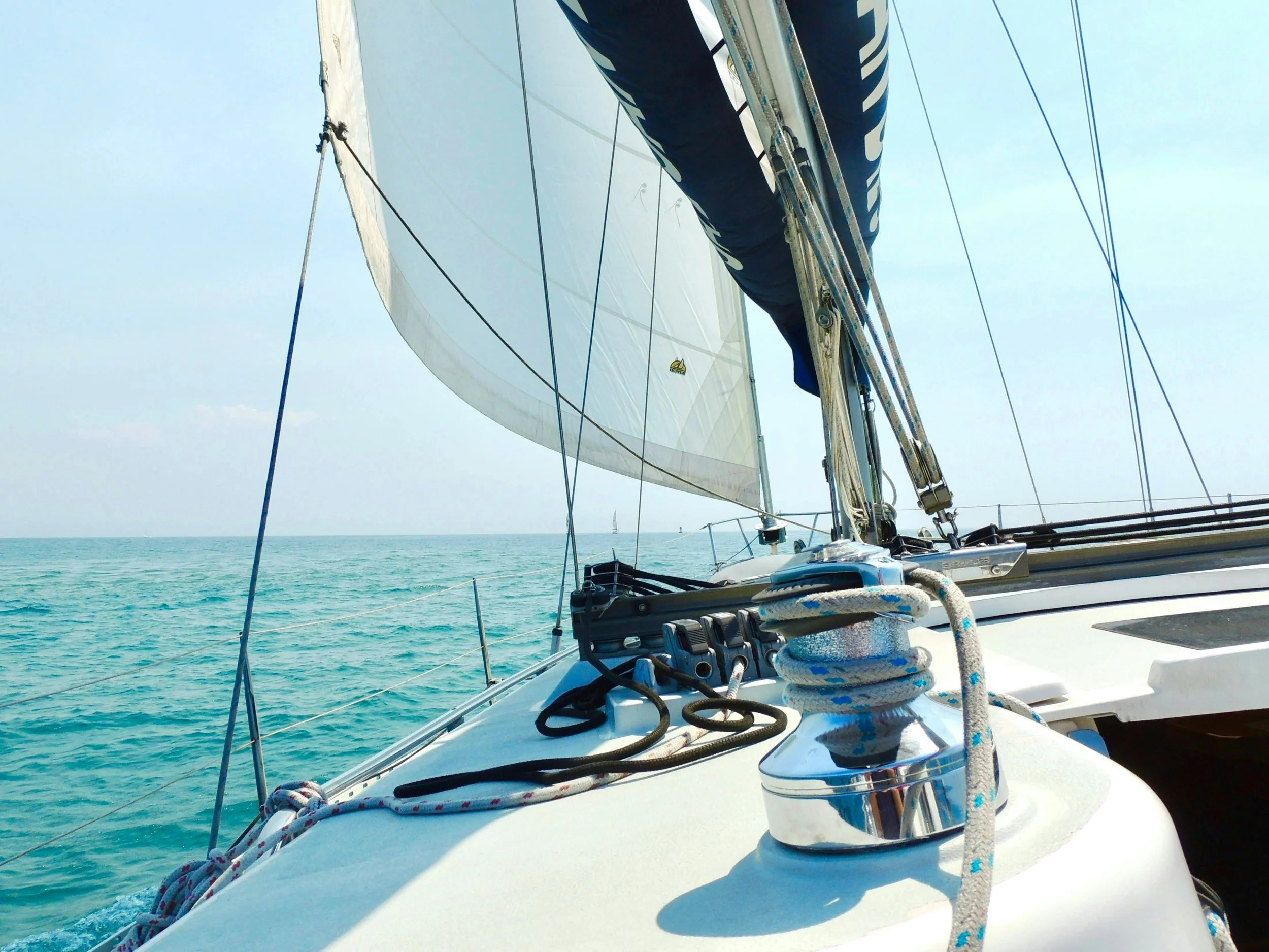 View from the bow of a sailboat showing the deck, ropes, and winch with the ocean and sky in the background.