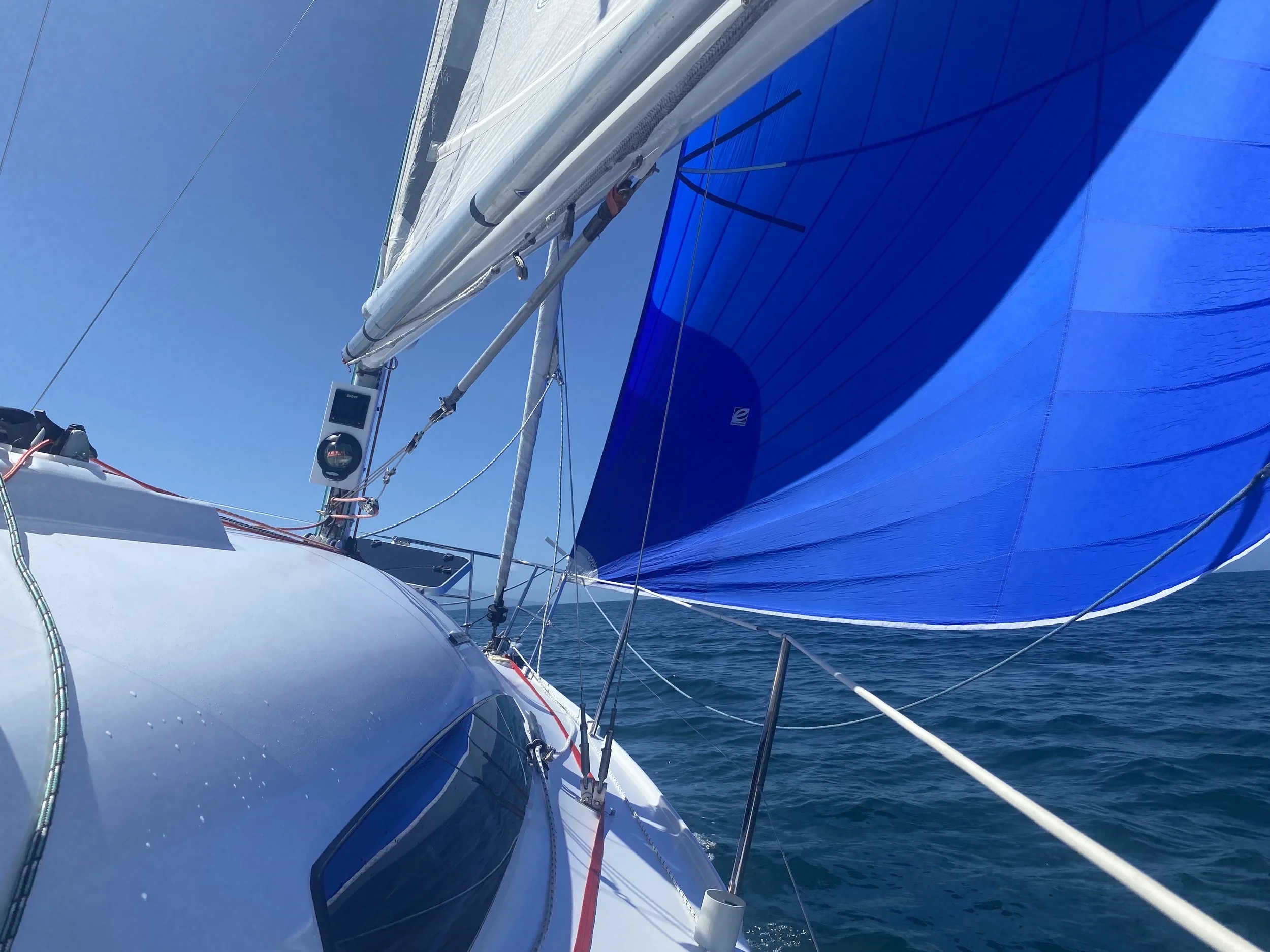 Looking up from the boat deck at the sailboat's rigging and bright blue spinnaker sail against a clear blue sky and ocean.