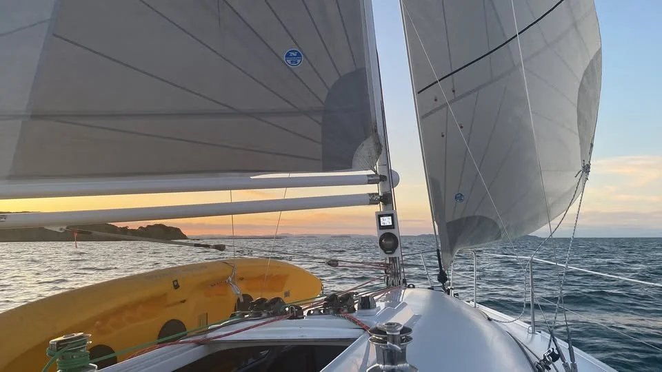 View from a sailboat at sunset showing the boat's deck, sails, and the ocean with land in the distance.