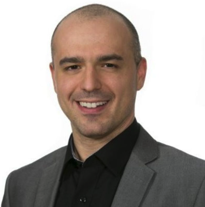 A man with a shaved head, smiling, wearing a dark suit jacket and black shirt, against a plain white background.