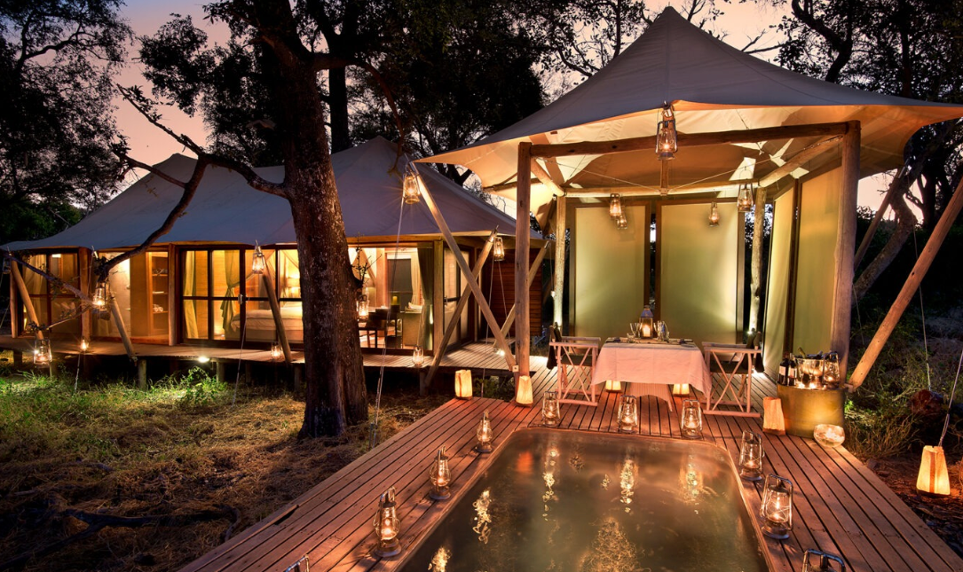 Candlelit luxury tented safari camp at dusk with a private dining deck, lanterns reflected in a plunge pool, and a glowing en-suite bedroom tent set among trees in the African bush