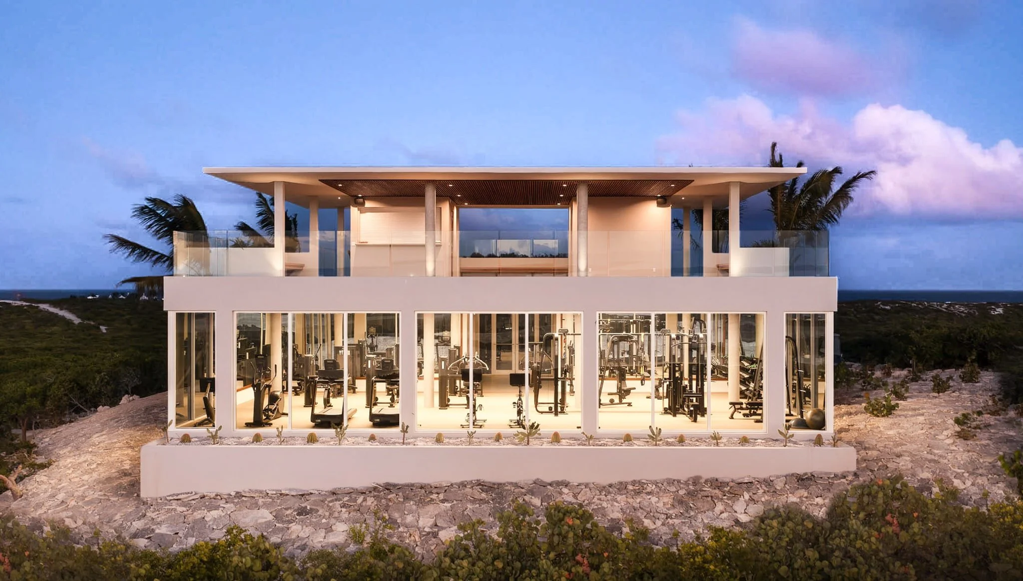 Glass-walled fitness center at Ambergris Cay in Turks and Caicos, lit from within at dusk and set on a rocky rise above the coastline