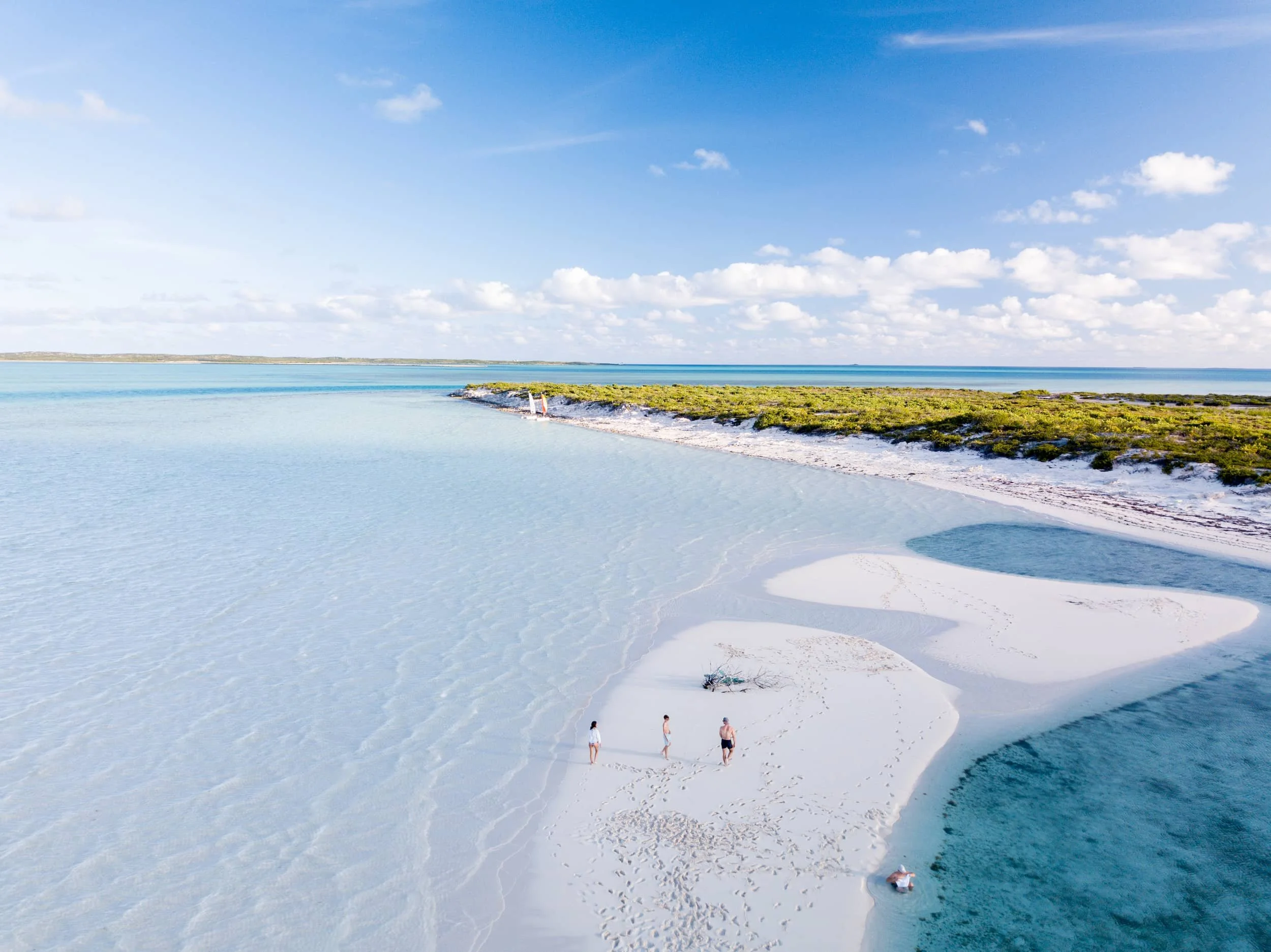 Three guests walking a deserted white sandbar at Ambergris Cay in Turks and Caicos, surrounded by shallow clear water with a small sailboat moored near the shoreline