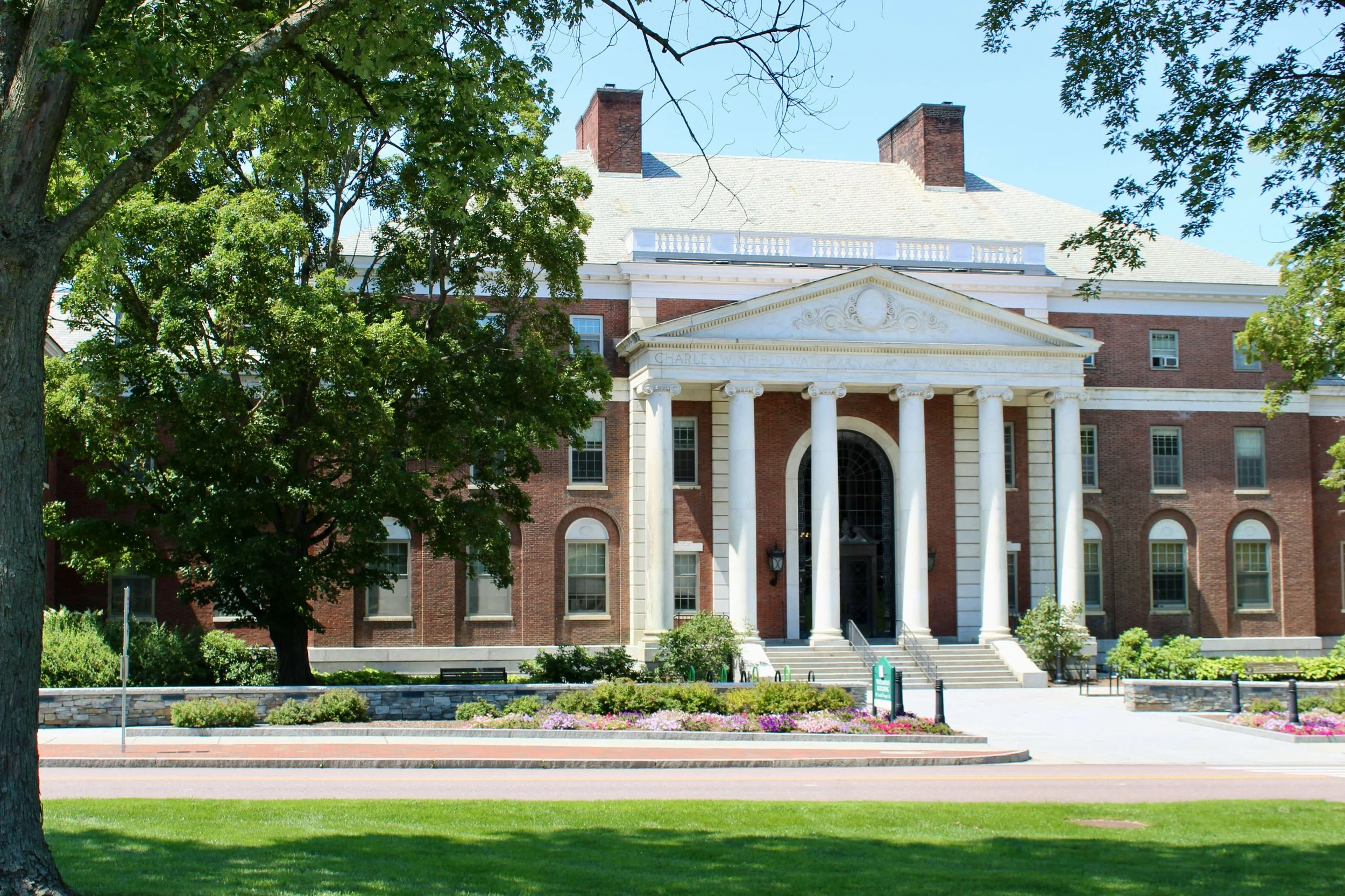 A historic brick building with white columns and a pediment, surrounded by trees and landscaping, on a sunny day.