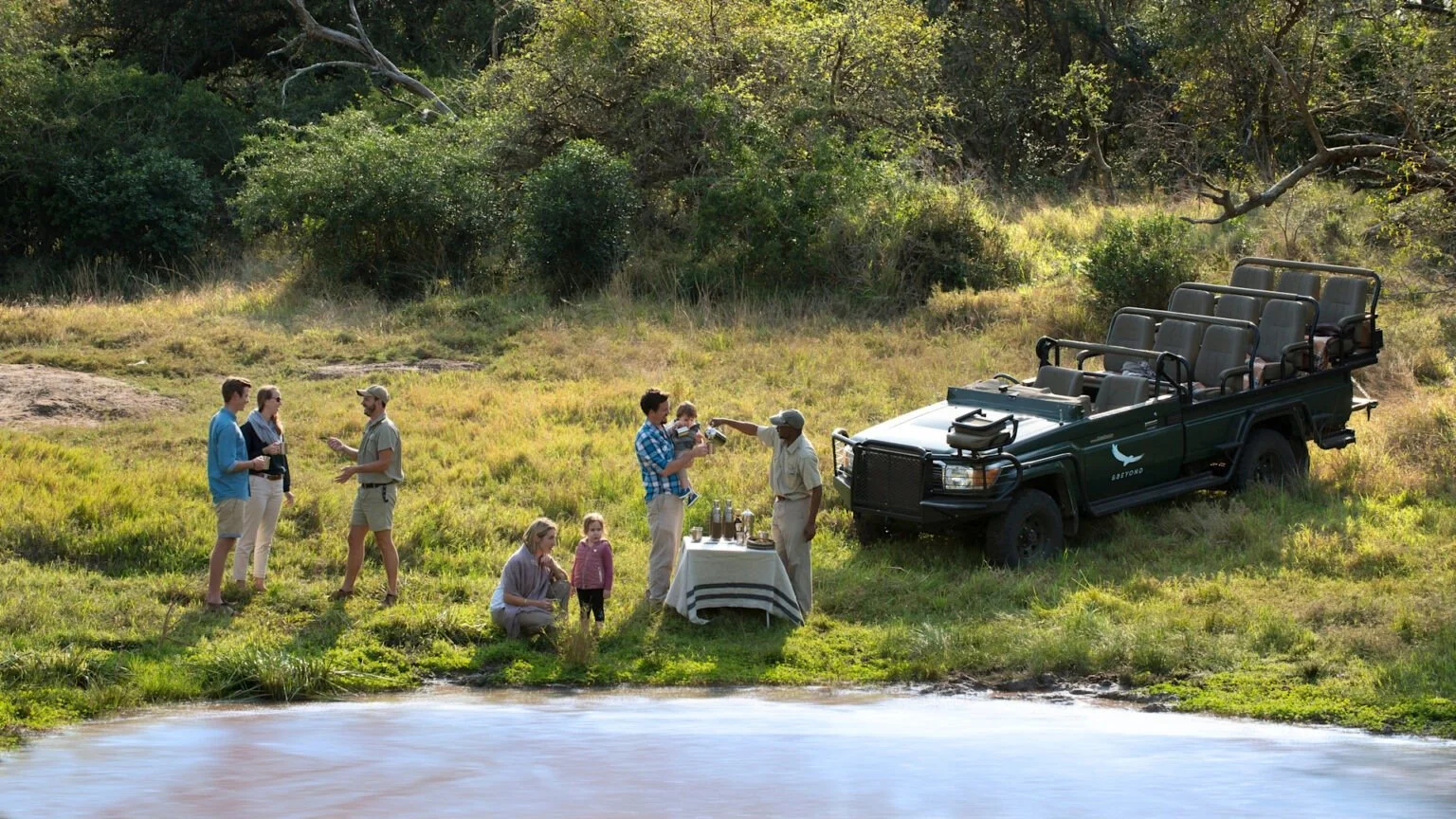 A family group enjoying a bush sundowner stop beside a river on a private game drive, with a guide serving drinks from a linen-covered table next to an open safari vehicle in the African bush