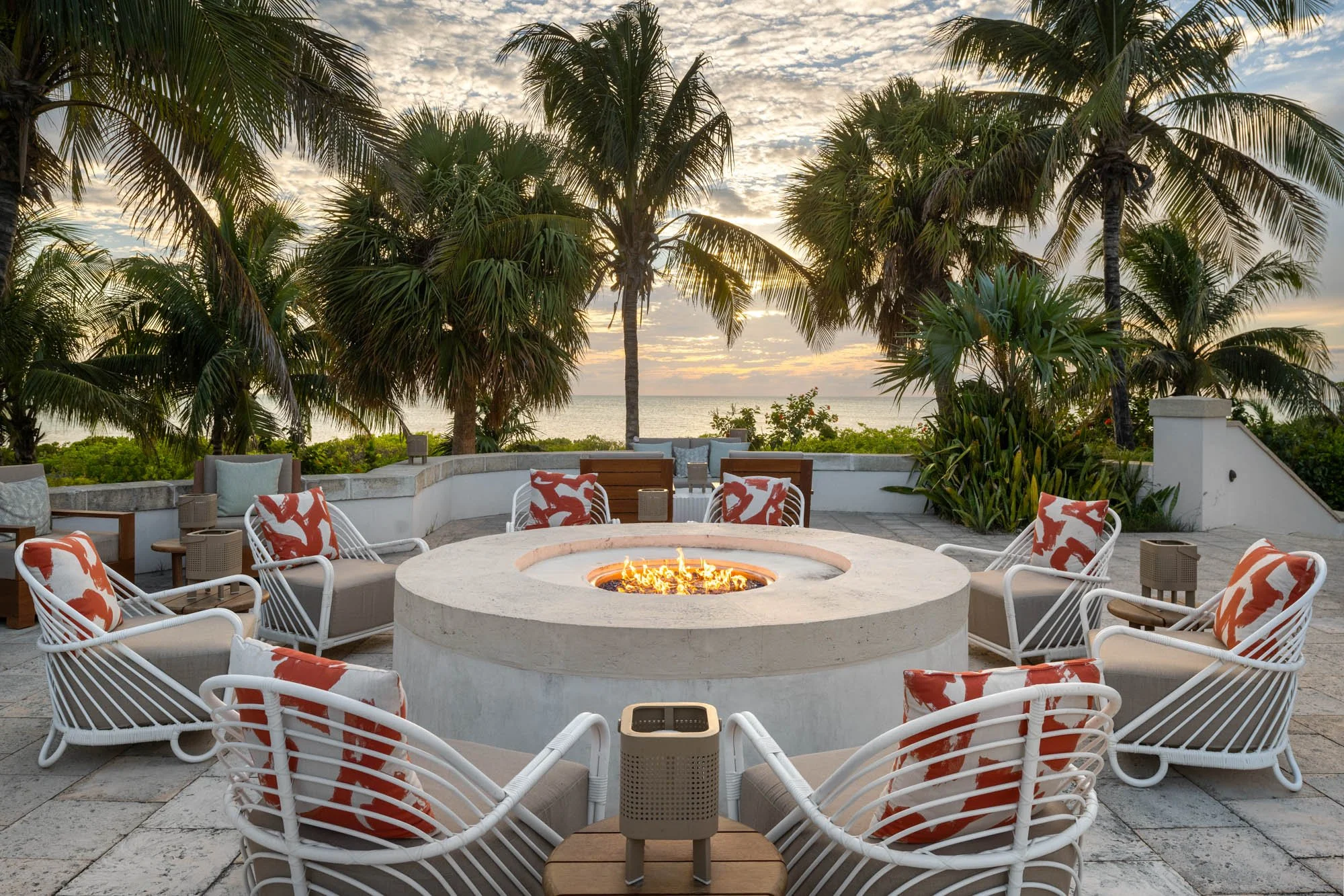 Circular oceanfront fire pit at Ambergris Cay in Turks and Caicos, ringed by white lounge chairs with coral cushions and framed by palms at sunset