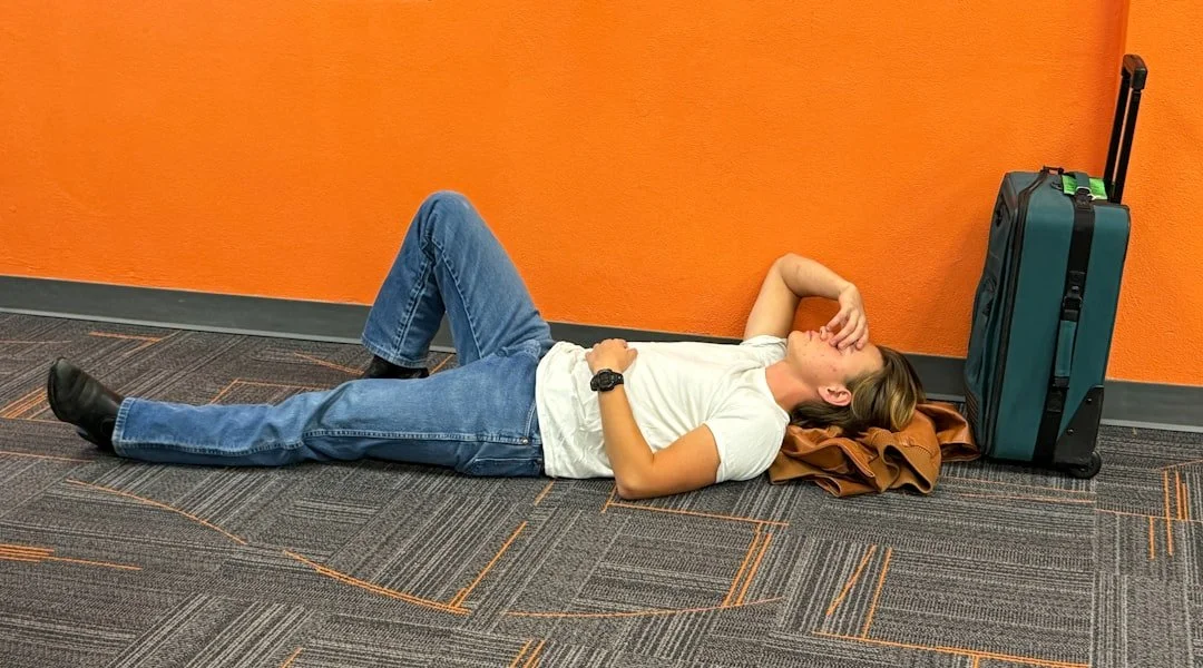 Tired traveler lying on airport floor next to rolling suitcase against orange wall