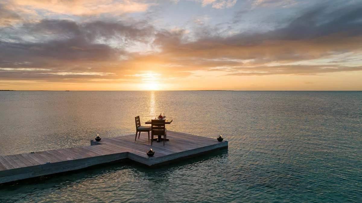 Private dinner for two set on a wooden dock over the water at Ambergris Cay in Turks and Caicos, lit by torches at sunset
