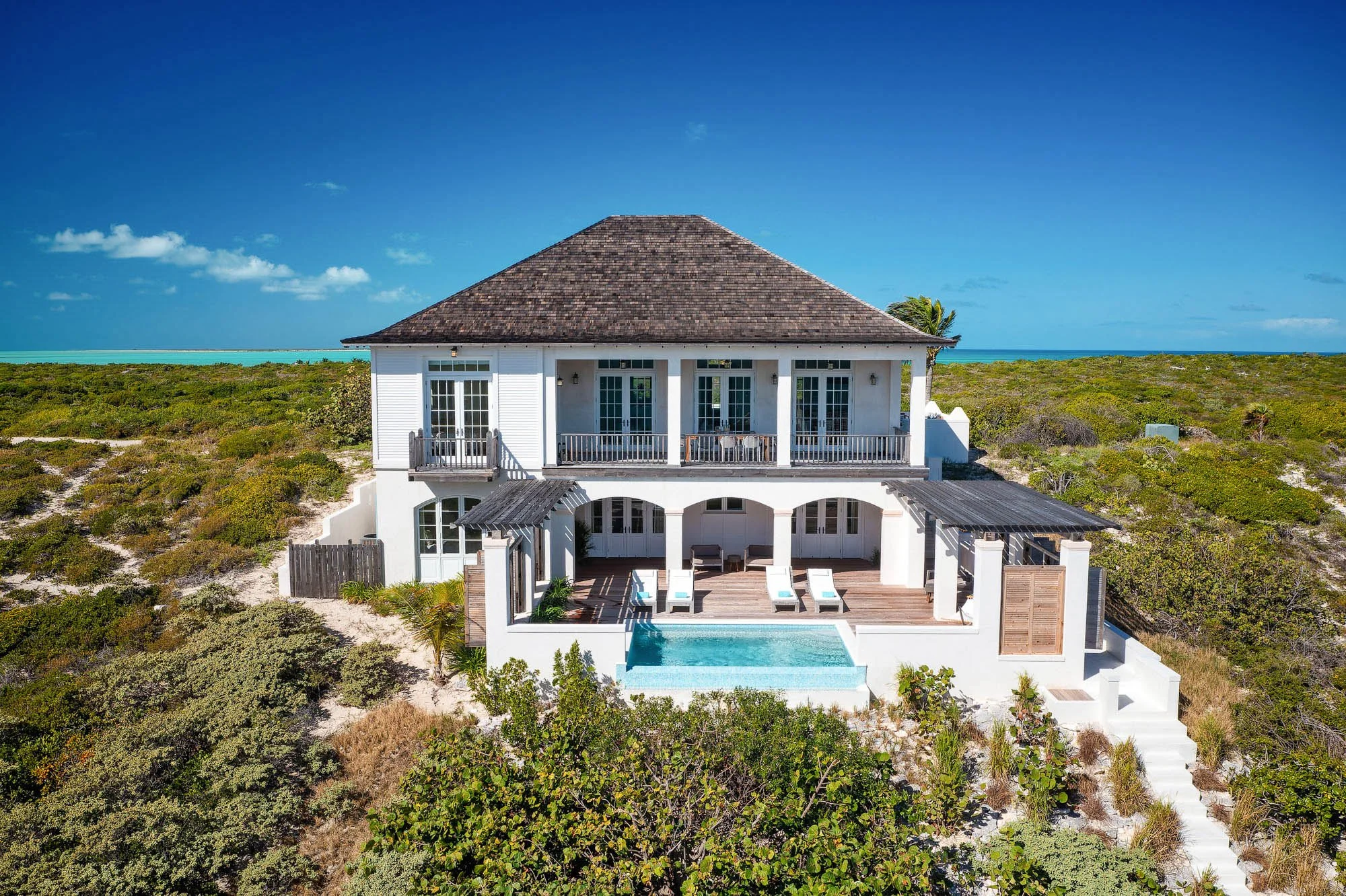 Two-story oceanview villa at Ambergris Cay in Turks and Caicos, with a private pool, shaded deck, and covered verandas overlooking low coastal brush and the sea