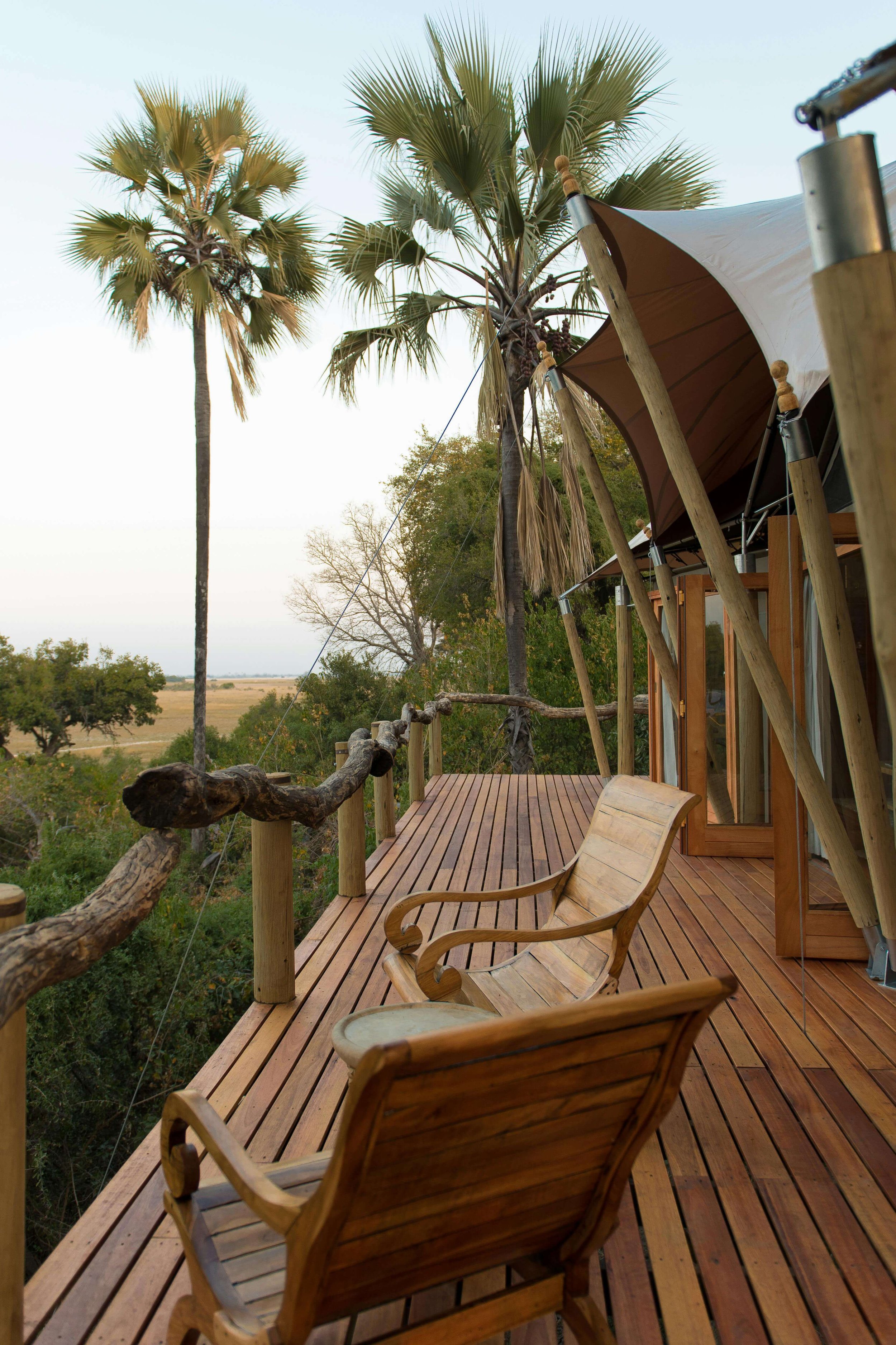 A wooden deck with two wooden chairs, surrounded by tropical palm trees and overlooking a distant landscape.