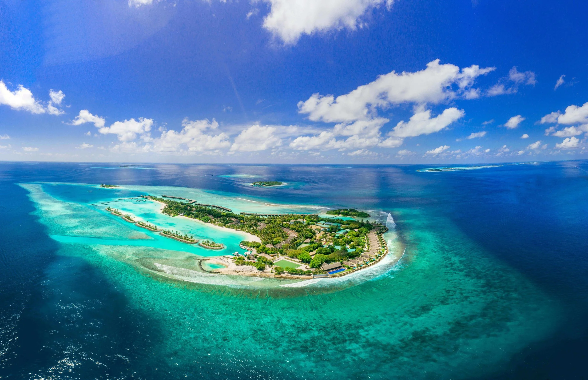 Aerial view of a lush island resort surrounded by turquoise ocean waters with white sandy beaches and coral reefs, under a partly cloudy sky.
