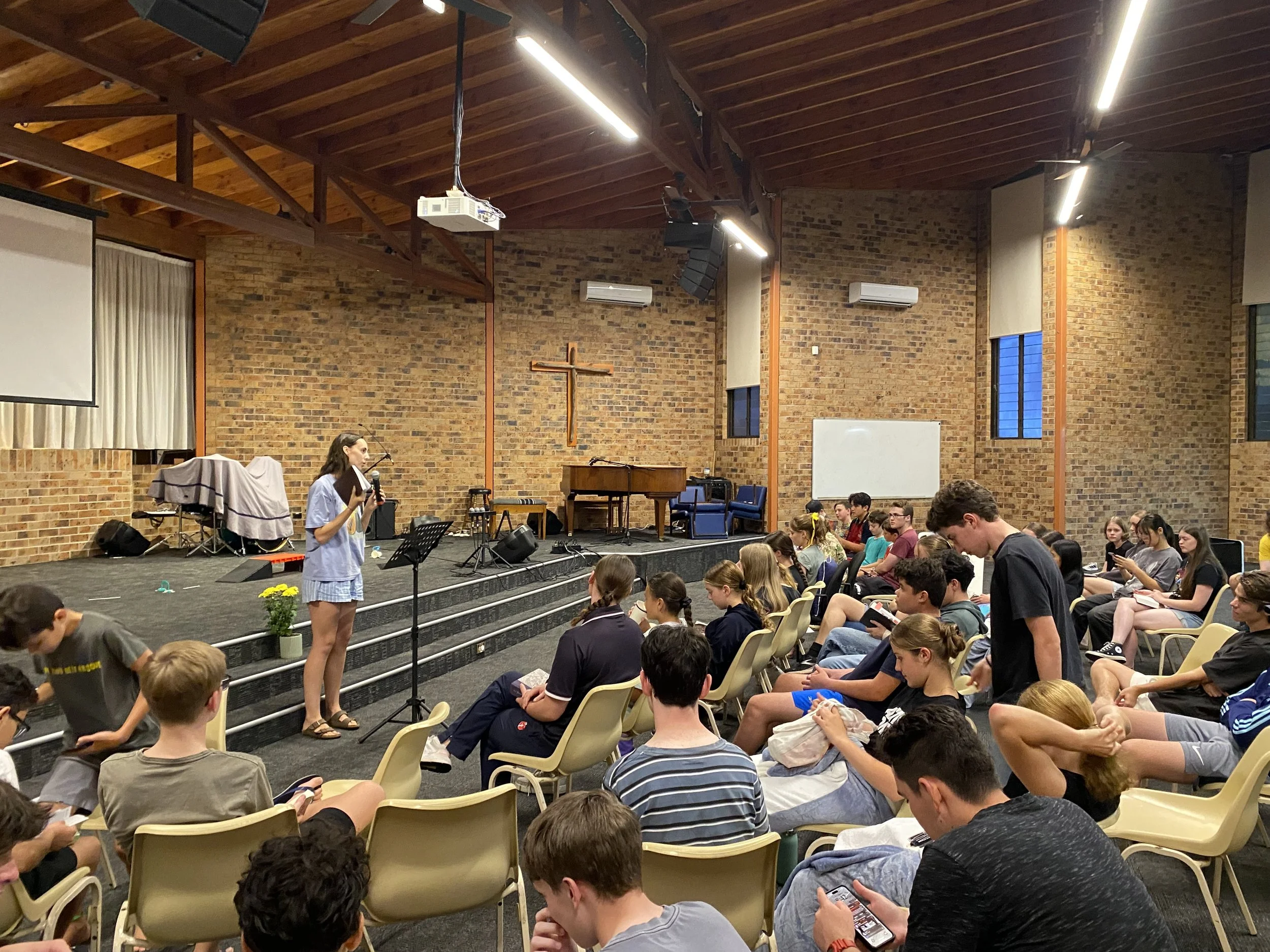 A young woman speaking into a microphone on a stage in front of a group of young people seated in chairs inside a brick-walled church or auditorium with a cross on the wall and musical instruments on the stage.