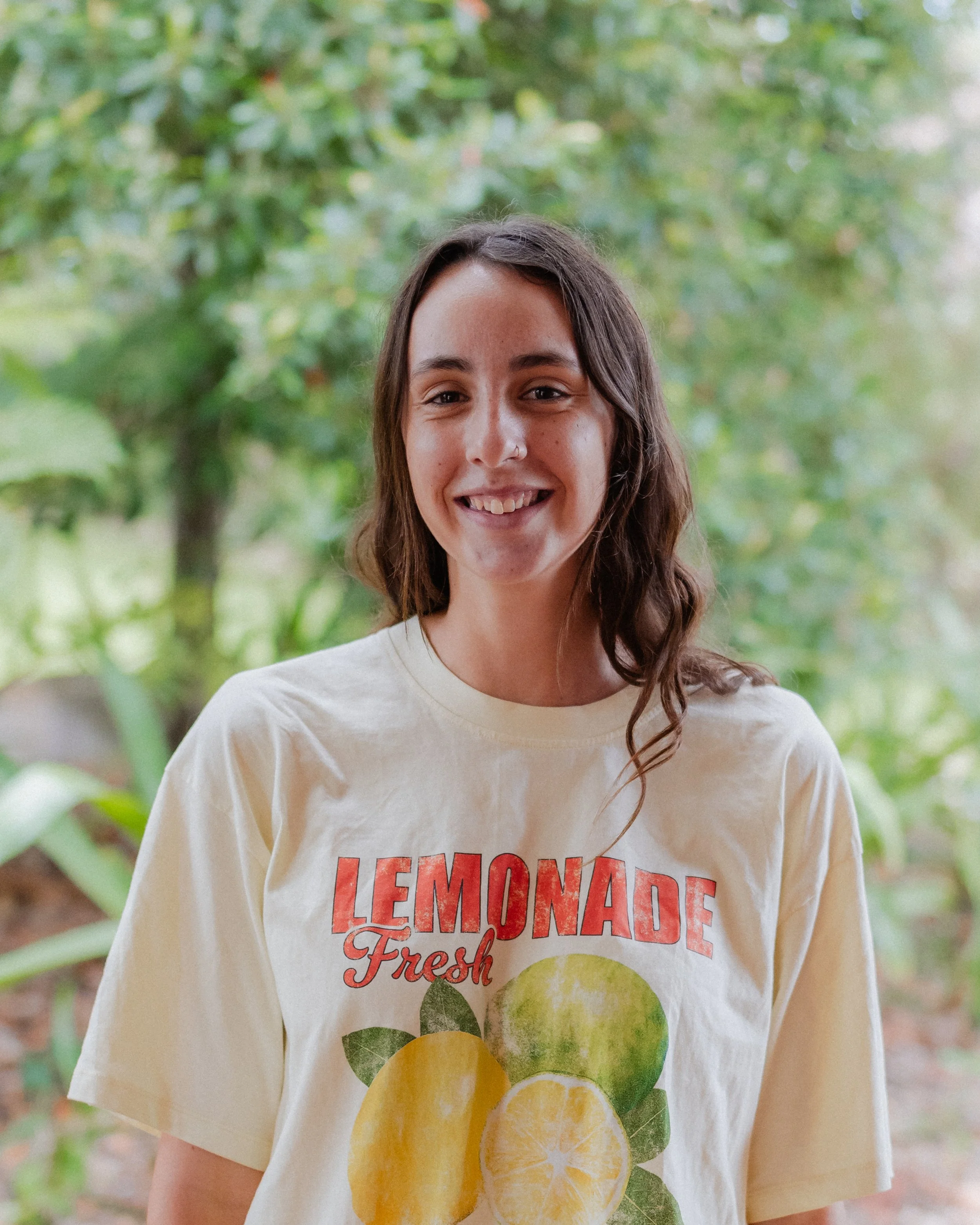 Young woman smiling outdoors, wearing a cream T-shirt with lemons and the words 'Lemonade Fresh' printed on it, with greenery in the background.