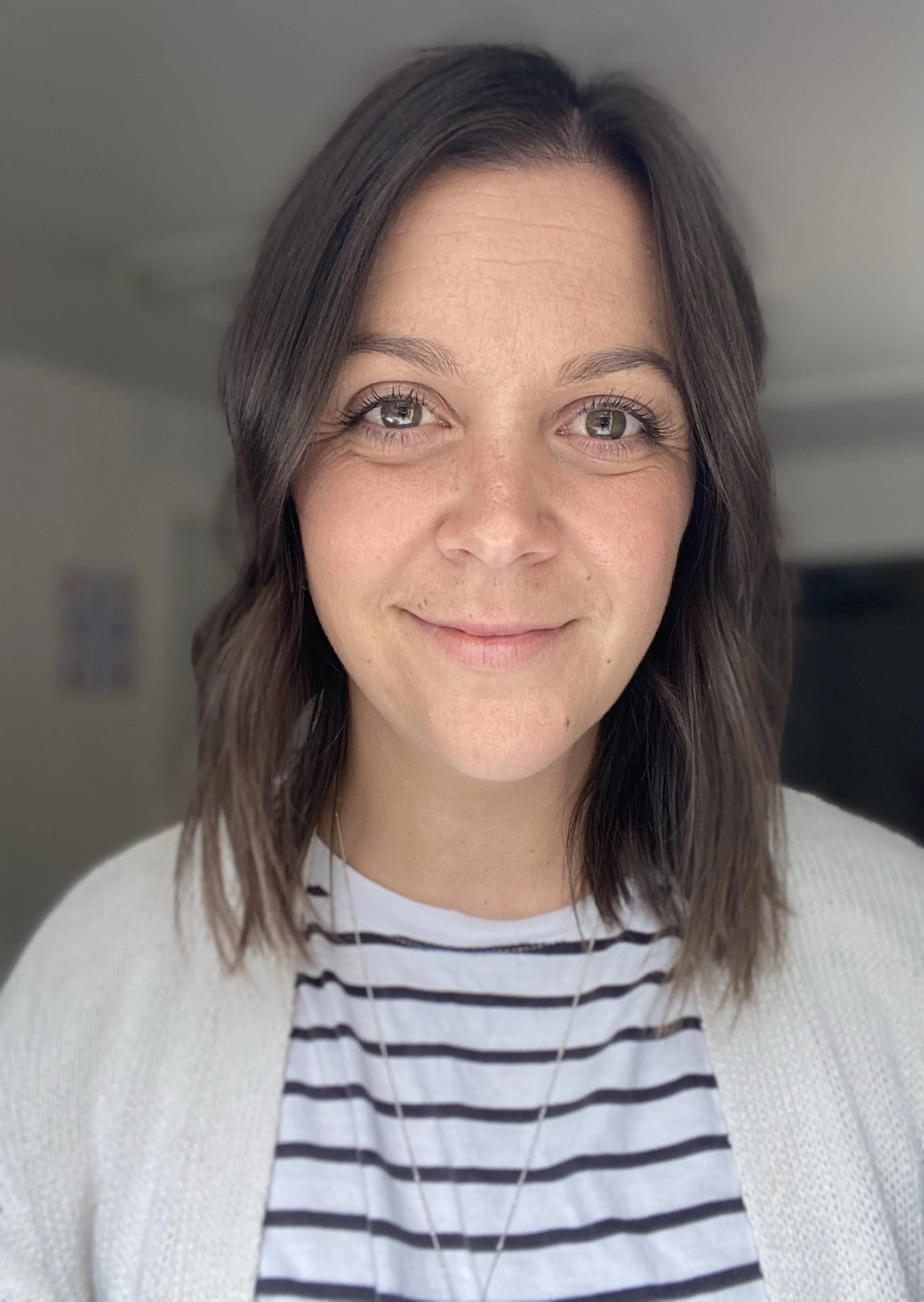 Close-up photo of a woman with shoulder-length brown hair, hazel eyes, and a slight smile, wearing a white cardigan over a black and white striped shirt.