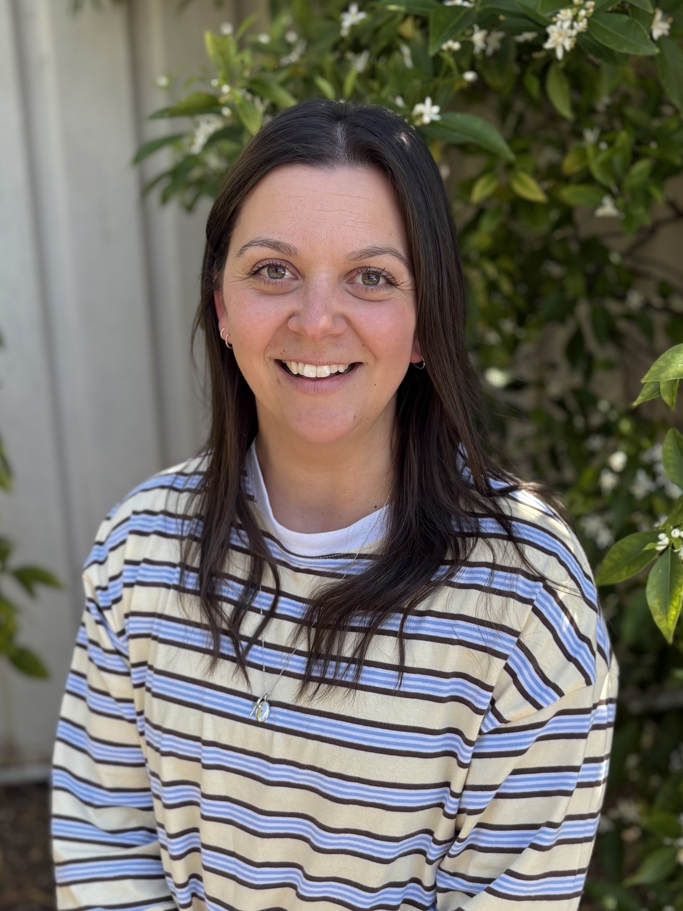 Close-up photo of a woman with shoulder-length brown hair, hazel eyes, and a slight smile, wearing a white cardigan over a black and white striped shirt.