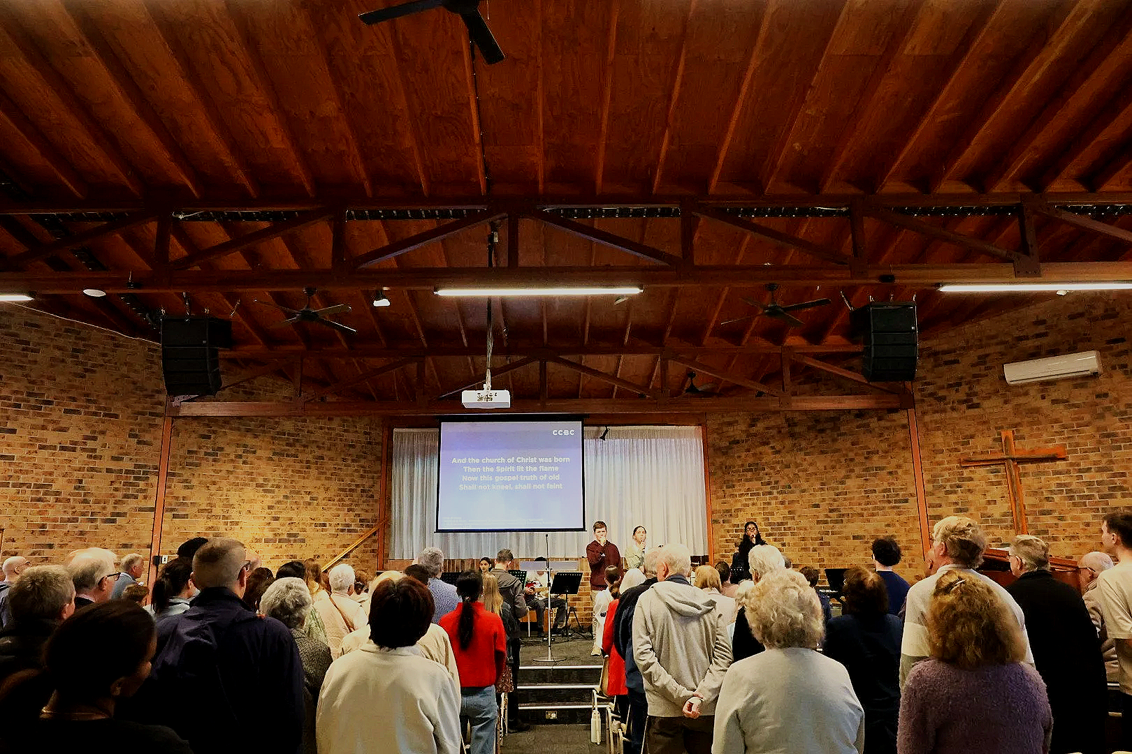 People gathered inside a church with a brick wall and wooden ceiling, listening to a speaker on stage with a large screen displaying text, and a cross on the wall.