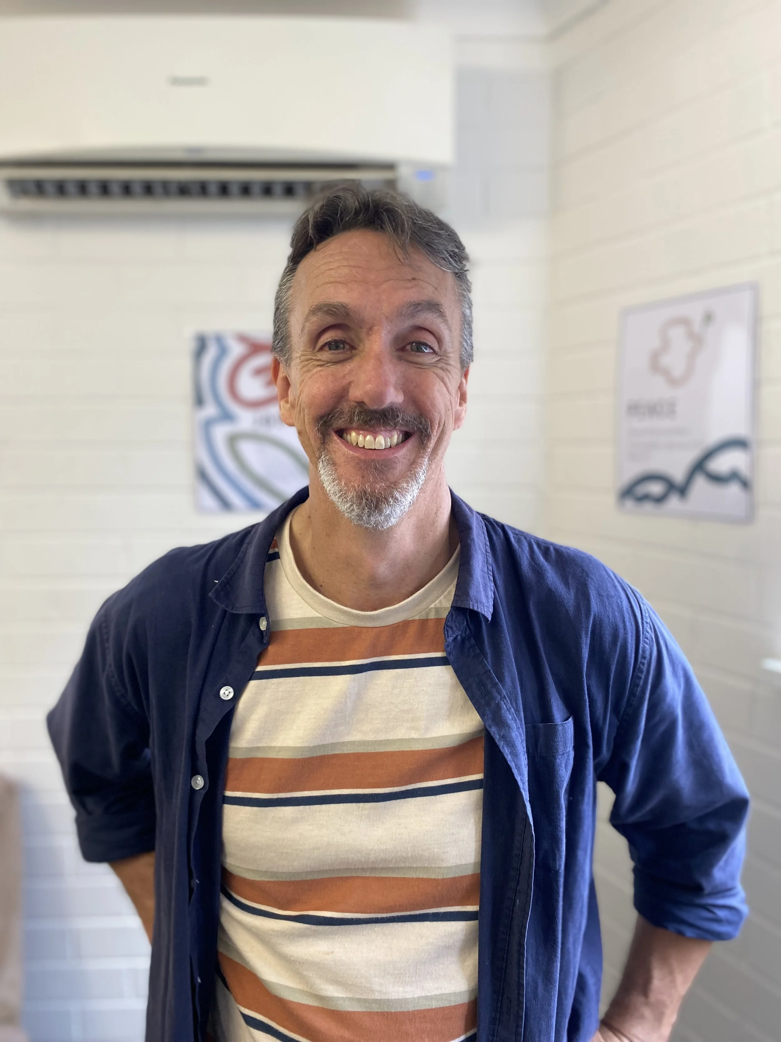 Smiling man with gray hair and beard, wearing a striped shirt and blue shirt, standing indoors with white walls and framed artwork in the background.