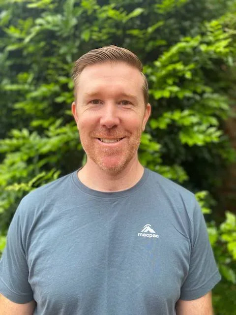 A man smiling outdoors with green foliage in the background, wearing a gray t-shirt with a small logo on the chest.