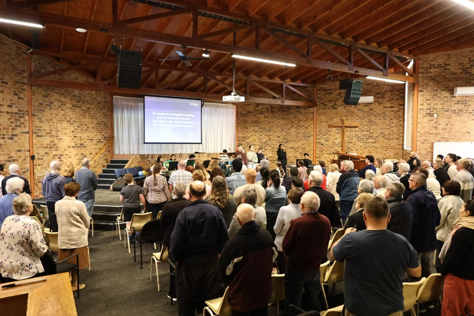 A large congregation of people standing in a church service with a brick wall interior, a cross, a stage with musicians, and a projector screen with lyrics.
